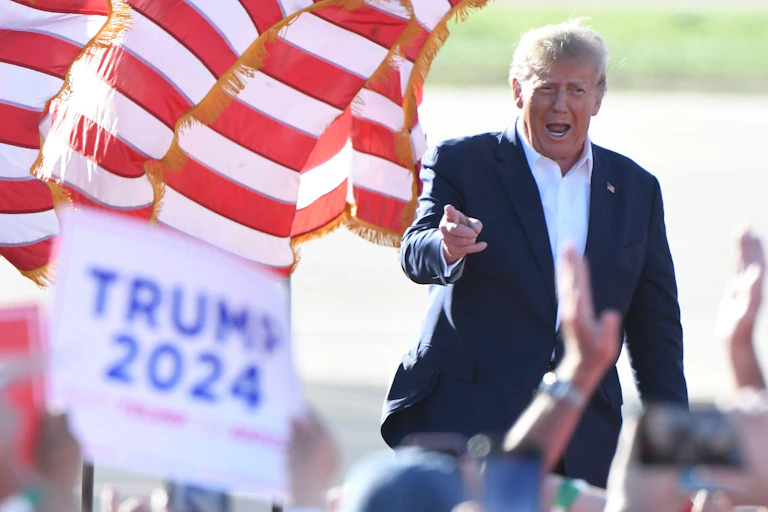 Donald Trump wearing suit jacket and white shirt in front of red and white striped banner.
