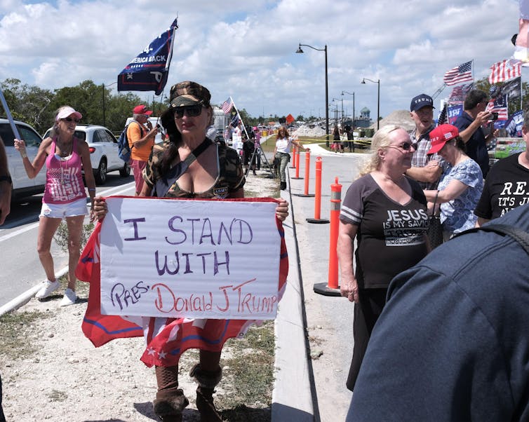 A woman in a black cap with a sign saying 'I stand with Pres. Donald J Trump'