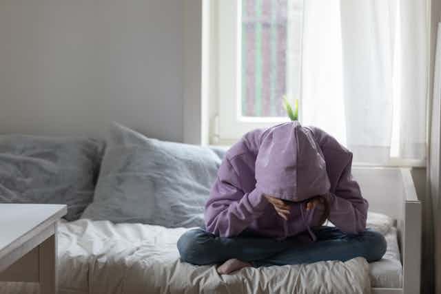 A young person wearing a hoodie sitting on a bed hunched over.