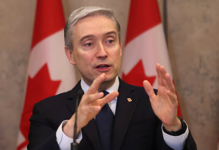 A white middle-aged man gestures with his hands while speaking. Two Canadian flags stand behind him.