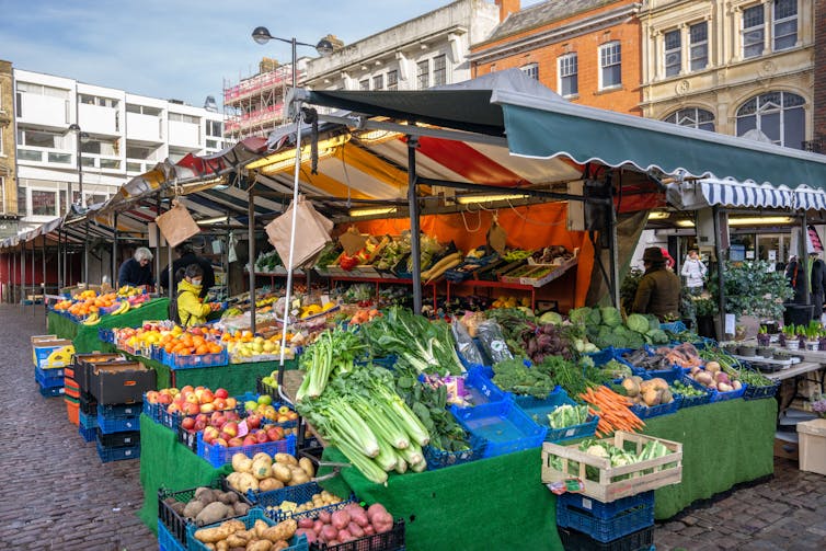 A greengrocer's fruit and vegetable stall, buildings and some shoppers in the background.
