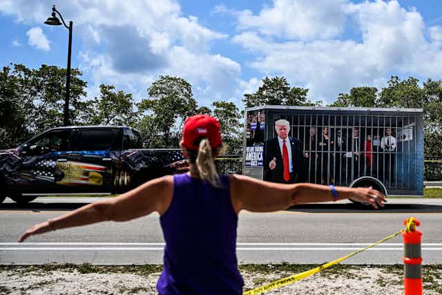 A woman spreads both of her arms out and faces a pick up truck that has a trailer that appears to be a jail cell, with a man wearing a dark suit and blonde hair outside of it, pointing his finger. 