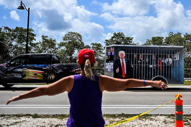 A woman spreads both of her arms out and faces a pick up truck that has a trailer that appears to be a jail cell, with a man wearing a dark suit and blonde hair outside of it, pointing his finger.