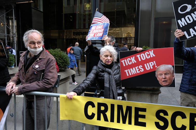 A woman with white hair holds a sign that says 'Tick tock, time's up' with the photo of a man's head on it. She and another few people stand behind a police barricade that has yellow tape on it and says 'crime scene.'