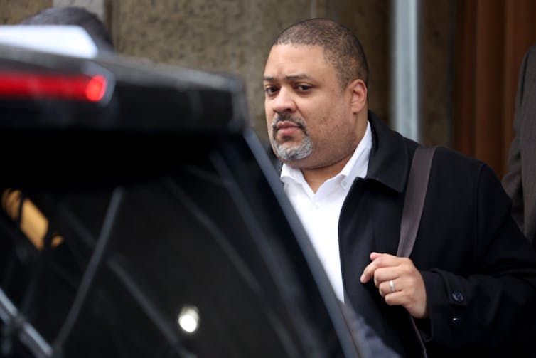 A Black man with a goatee wears a dark coat, white shirt and appears to walk toward a waiting car.
