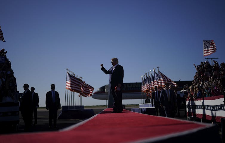 A man is shown in silhouette raising his fist and standing on a red carpet surrounded by American flags.