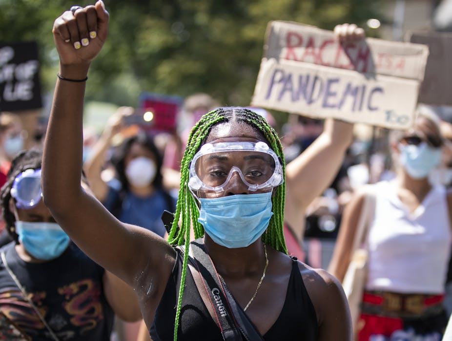 A black woman wearing goggles and a mask raises her fist in the air. 