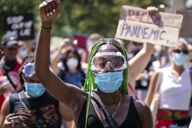 A black woman wearing goggles and a mask raises her fist in the air.