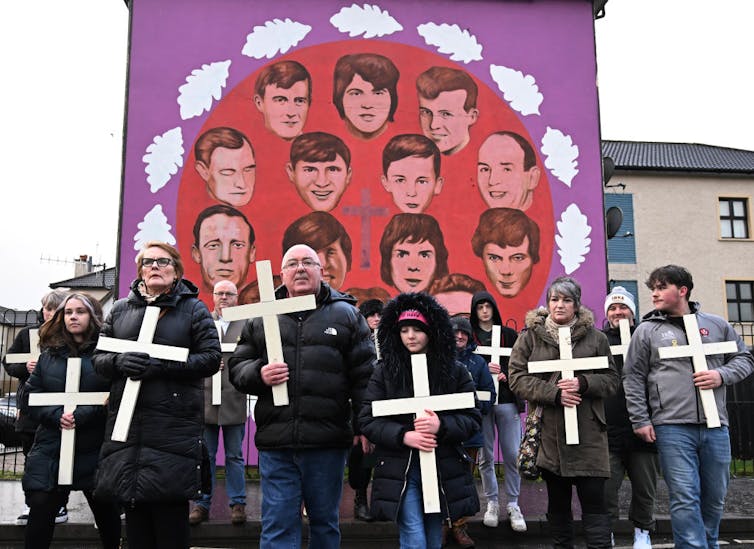 People in dark coats hold white crosses in front of a purple and red mural with people's faces painted in it.