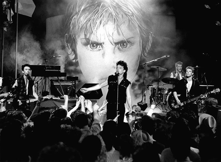 A black and white photo shows a band performing on stage in front of a large illustration of a boy's face.