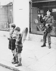 A soldier stands on a street as two young children, one holding a fake shield, stand in front.