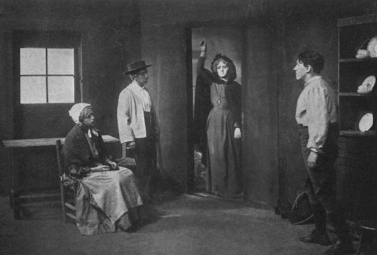 A black and white picture of a woman holding up a lantern in a doorway to a room with three people in it.