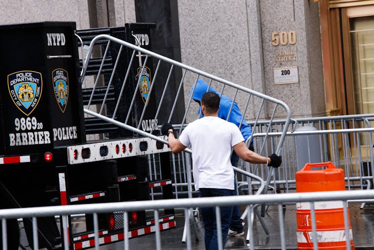 A man in a a white T-shirt setting up a metal barricade in front of a building.