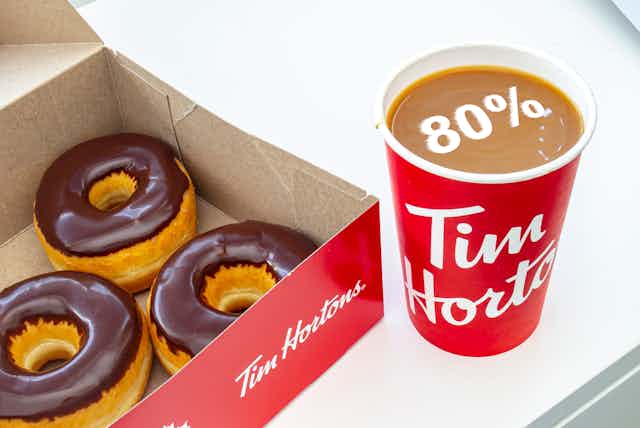 A picture of a Tim Hortons coffee cup with 80 % displayed floating in the cup, which is beside a box of chocolate glazed donuts. 