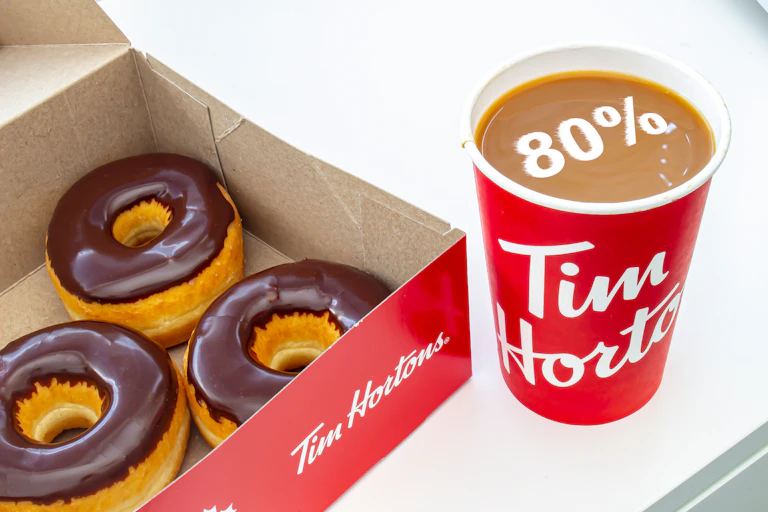 A picture of a Tim Hortons coffee cup with 80 % displayed floating in the cup, which is beside a box of chocolate glazed donuts.