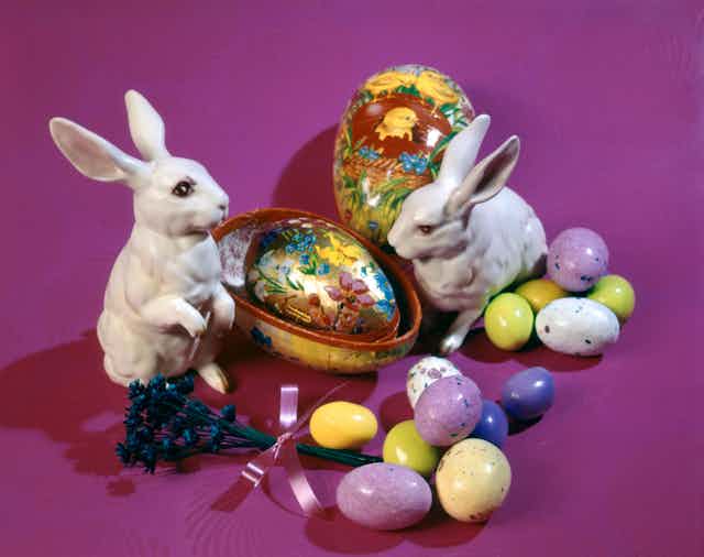 Two ceramic rabbits positioned among candy eggs and painted eggs against a fushia background.