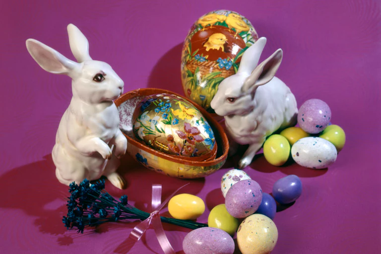 Two ceramic rabbits positioned among candy eggs and painted eggs against a fushia background.