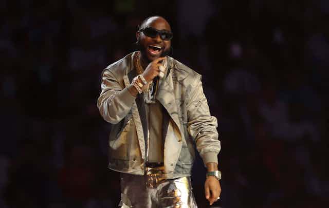 A man in sunglasses and a gold suit sings into a microphone against a black backdrop.