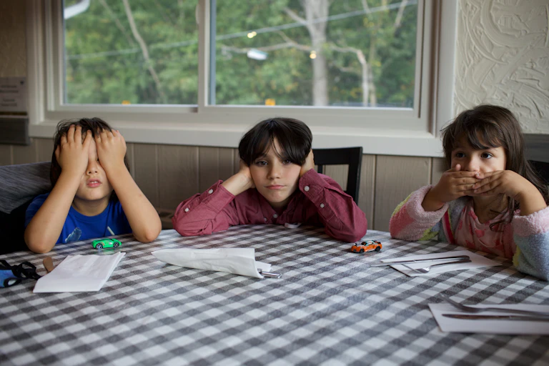 Three kids sitting at a table, each covering their eyes, ears and mouth.