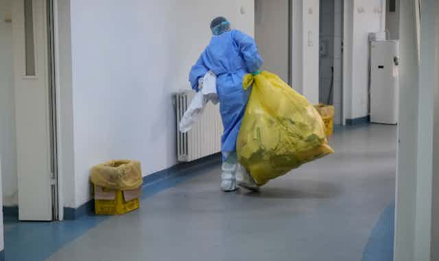 A person in blue personal protective equipment carries a large yellow plastic garbage bag down a hospital corridor