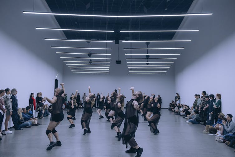 A white room, an audience around the edge, a mass of dancers in black.