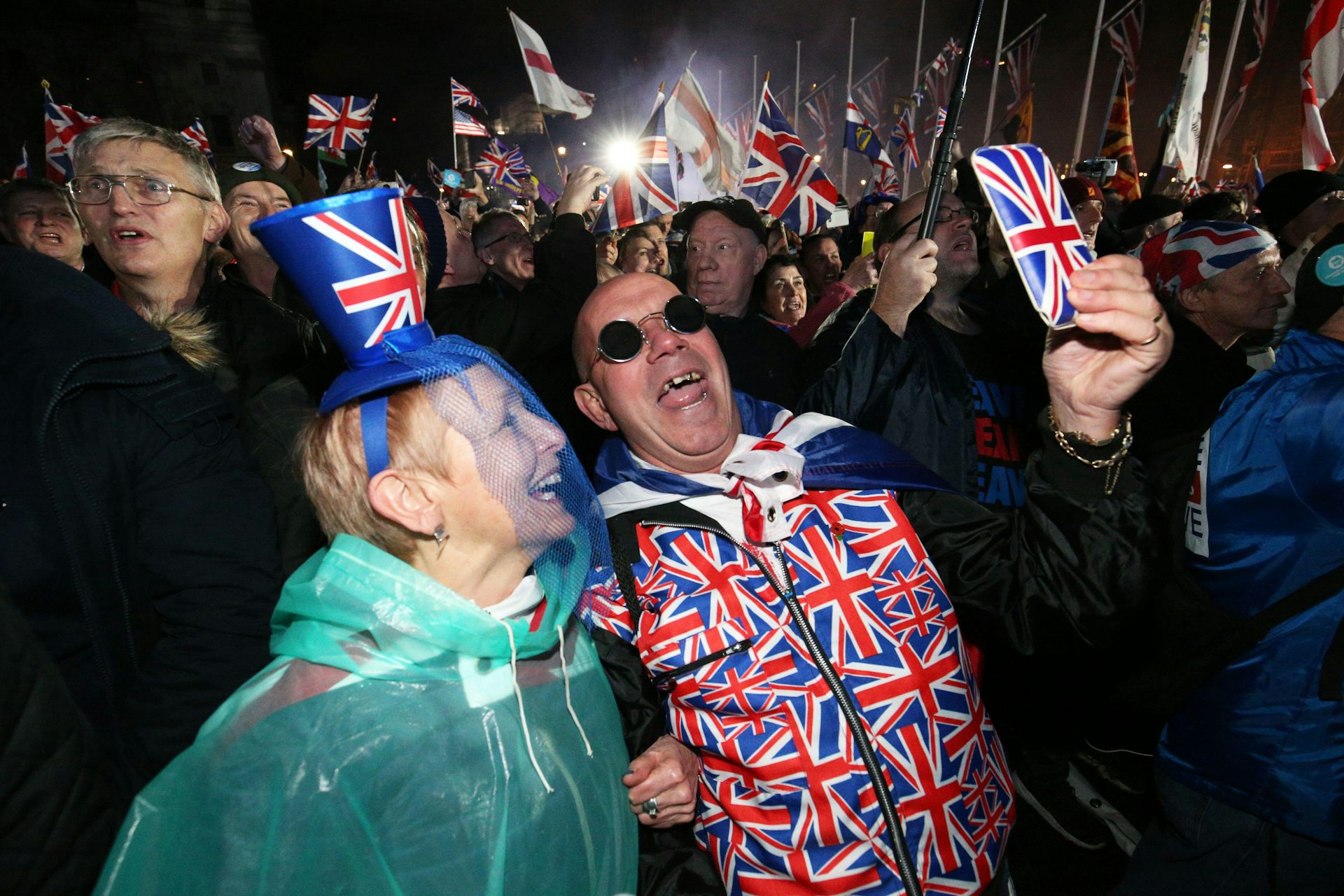 A woman and a man wearing union flag clothing take a selfie using a phone in a union flag case amid a crowd of people waving union flags.