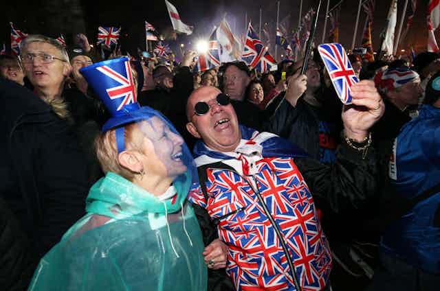 A woman and a man wearing union flag clothing take a selfie using a phone in a union flag case amid a crowd of people waving union flags.
