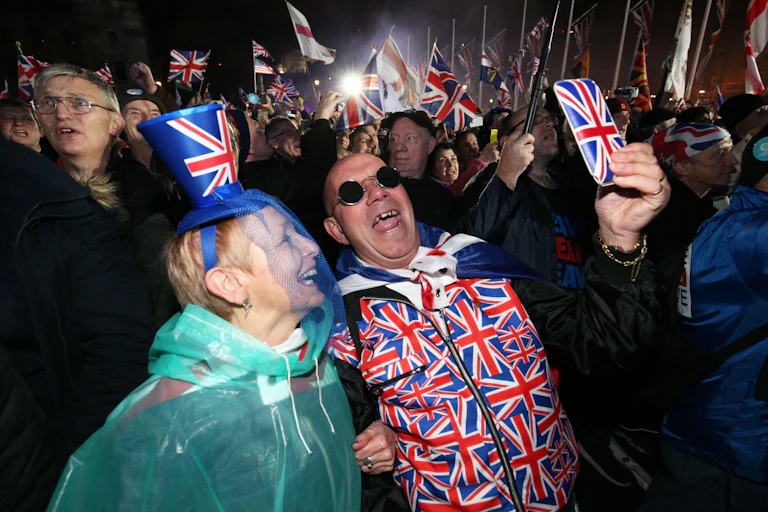 A woman and a man wearing union flag clothing take a selfie using a phone in a union flag case amid a crowd of people waving union flags.