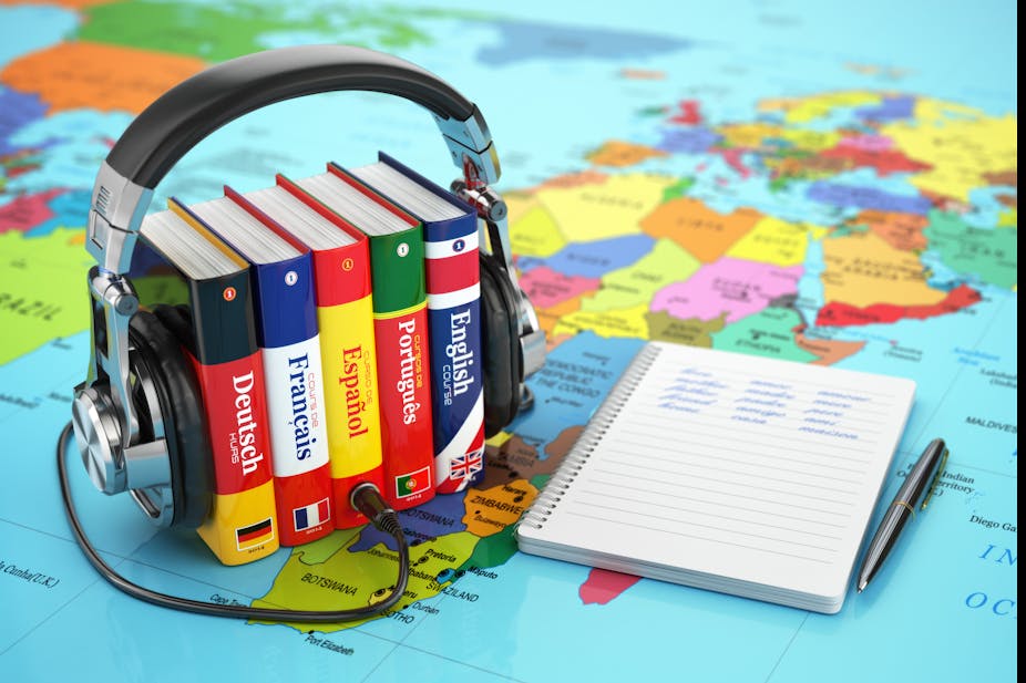 A line of European language dictionaries wears a set of headphones. Next to it is a notepad and pen. In the background is a map of the world.
