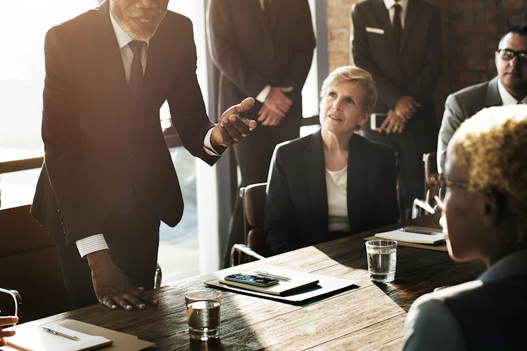 A business meeting with a diverse group of adults in professional clothing, around a table in an office. One man is standing up and gesturing with his hands as he talks.