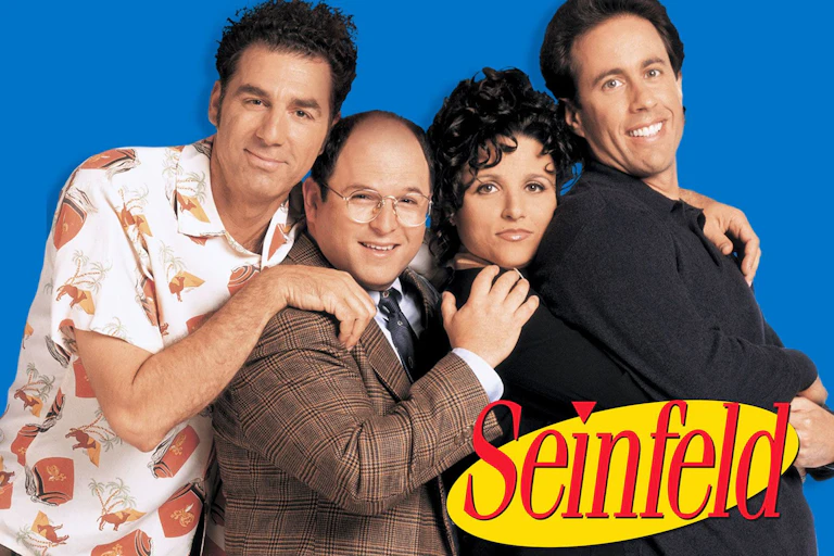 Four people stand looking towards us shoulders forward. The man on the left wears a whacky shirt, while the man next to him wears a brown checked jacket. The woman wears black and the man on the right hand side wears a navy blue shirt. All are smiling to varying degrees