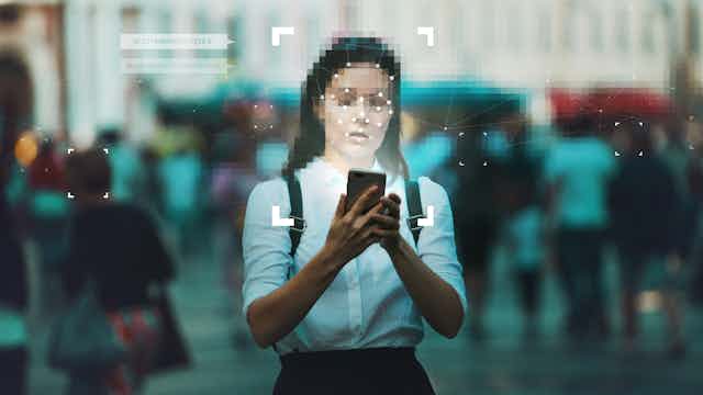 a woman on a city street holds a phone, her face is pixelated