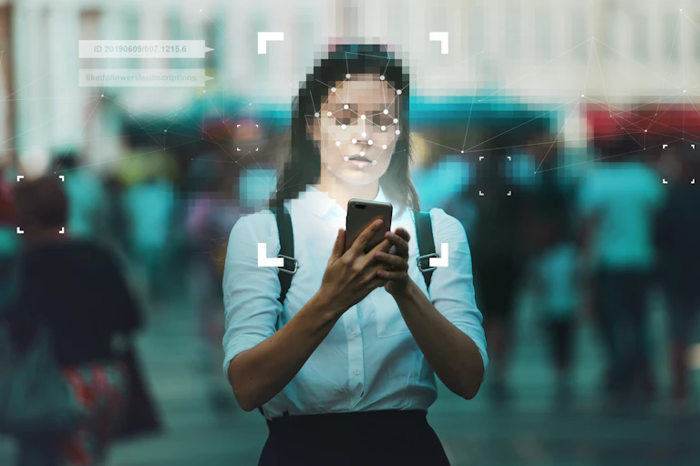 a woman on a city street holds a phone, her face is pixelated
