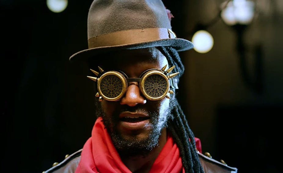 A man with dreadlocks and a hat wears futuristic glasses.