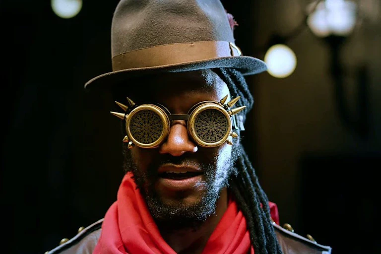 A man with dreadlocks and a hat wears futuristic glasses.