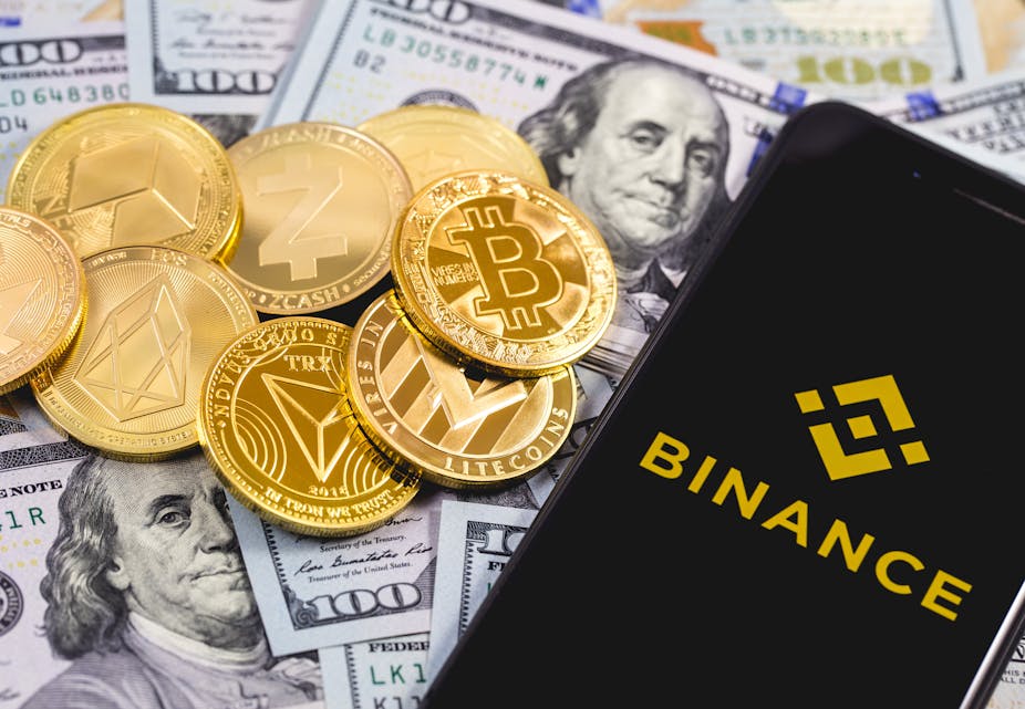 Apple iPhone with Binance logo (gold on black), background of dollars with a pile of cryptocurrency coins.