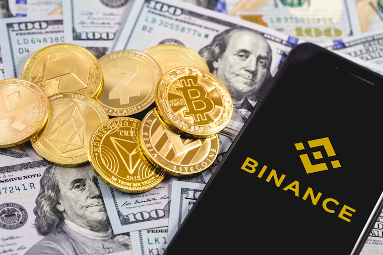 Apple iPhone with Binance logo (gold on black), background of dollars with a pile of cryptocurrency coins.