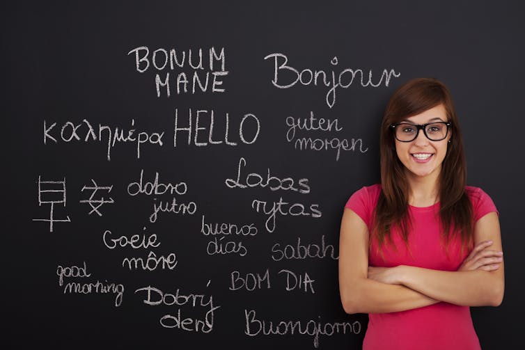 A woman wearing a pink top stands with arms crossed in front of a chalkboard, which features a range of words in different languages which mean 