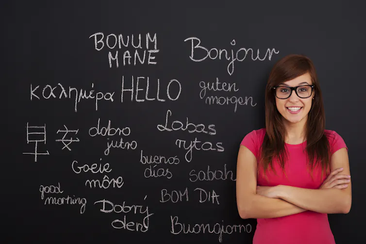 A woman wearing a pink top stands with arms crossed in front of a chalkboard, which features a range of words in different languages which mean