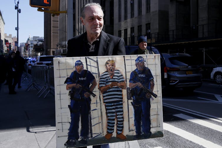 An older looking white man holds up a painting that shows a man wearing black and white striped prison uniform, flanked by two other men.