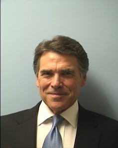 A man with dark hair and a dark suit smiles. He stands against a plain gray background.