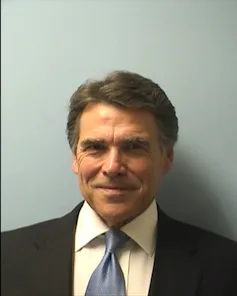 A man with dark hair and a dark suit smiles. He stands against a plain gray background.