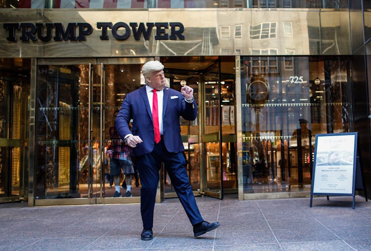 A person with a rubber mask on looking like an old man with white hair and a blue suit does a dance outside of a building that says Trump Tower
