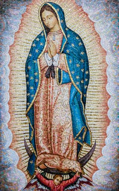A mosaic of Our Lady of Guadelupe, a virgin saint. She wears a long coral robe and blue starry hooded cape, hands clasped in prayer.