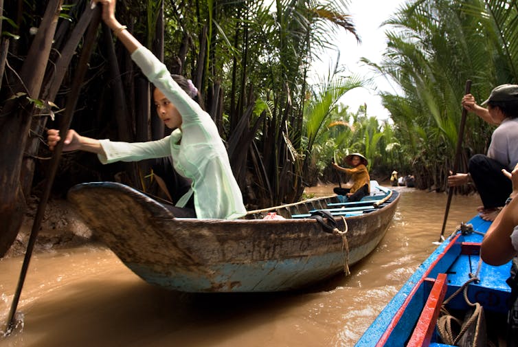 Two women row a small boat in through a narrow channel in the Mekong Delta. Another boat is passing them.