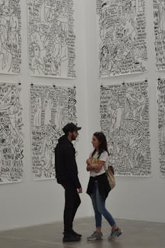 Two young people talk together next to giant black and white drawings pinned on the walls behind them.