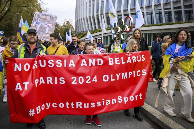 A crowd of people hold a banner that says 'No Russia and Belarus at Paris 2024 Olympics'