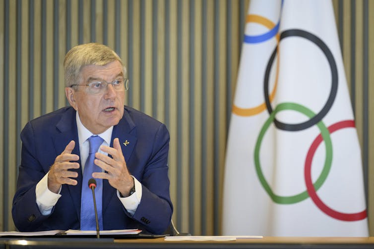 A white older man in a suit gestured while speaking from behind a desk. The Olympic flag stands in the background.