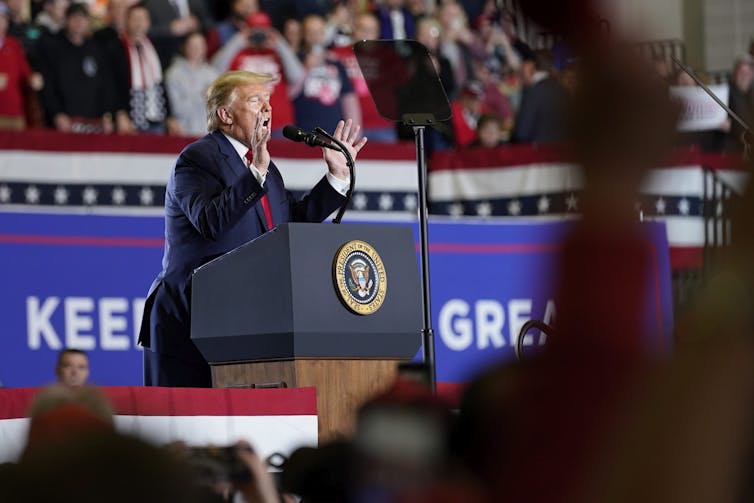 A rotund man with raised arms speaks into a microphone at a podium with the presidential seal on it.