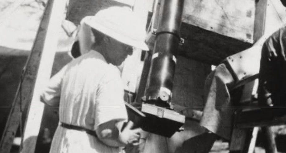 A woman in a white dress and hat operating a telescope on a wooden frame